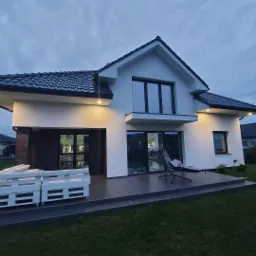 Nowoczesny dom jednorodzinny z białymi ścianami i ciemną dachówką, oświetlony zewnętrznymi lampami, z tarasem wyłożonym szarymi płytkami i meblami ogrodowymi. Widoczne elementy wykończenia elewacji...