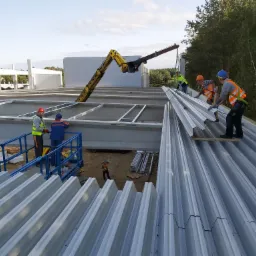 Montaż paneli dachowych na konstrukcji stalowej hali, z użyciem dźwigu i platform roboczych, widok z g&oacute;ry na ekipę w kaskach i kamizelkach odblaskowych.