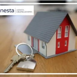 Mały model domu z czerwonym akcentem na drewnianym stole, z kluczami na pierwszym planie, logo agencji ubezpieczeniowej w lewym g&oacute;rnym rogu.