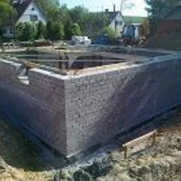 Fundamenty domu w trakcie murowania z bloczków betonowych, widoczne szalunki i zbrojenie, w tle sprzęt budowlany i drzewa.