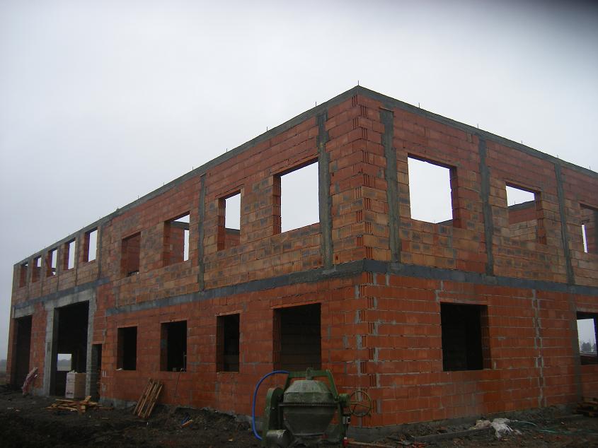 Surowy stan dwukondygnacyjnego budynku z czerwonej cegły z otworami okiennymi, widoczna betoniarka i palety pod budynkiem, pochmurne niebo w tle.