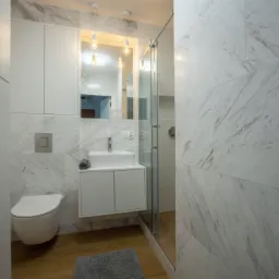Nowoczesna łazienka wykończona płytkami imitującymi biały marmur, z wiszącą toaletą, szafką pod umywalką i kabiną prysznicową ze szklanymi drzwiami.