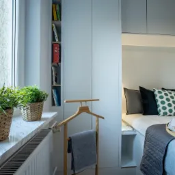 Jasny pokój z marmurowym parapetem, na którym stoją dwie plecione doniczki z zielonymi roślinami, oraz wnęką z łóżkiem i szafkami nad nim.