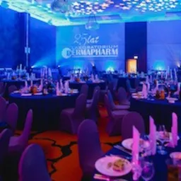 Sala bankietowa przygotowana na uroczystość firmową: okrągłe stoły z ciemnymi obrusami, elegancka zastawa, krzesła w pokrowcach, oświetlenie sceniczne w kolorze niebieskim i dekoracja świetlna...