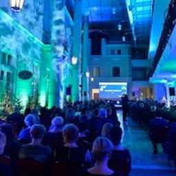 Agencja Eventowa Tajemnice Imprez - Wnętrze sali konferencyjnej oświetlone niebieskim i zielonym światłem, widoczny tłum ludzi siedzących na krzesłach i prelegent na scenie przed ekranem.