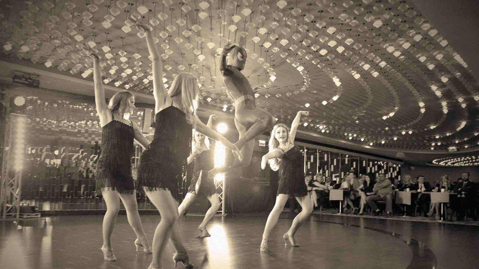 Czarno-białe zdjęcie grupy tancerek w czarnych sukienkach z frędzlami, prezentujących układ taneczny na scenie z publicznością w tle, z jednym tancerzem w stroju Spidermana wykonującym skok.