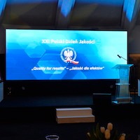 Scena z dużym ekranem LED wyświetlającym logo i napis 'XXI Polski Dzień Jakości - Quality for results - Jakość dla efektów'. Na pierwszym planie mównica i dekoracje.