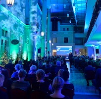 Wnętrze sali konferencyjnej oświetlone niebieskim i zielonym światłem, widoczny tłum ludzi siedzących na krzesłach i prelegent na scenie przed ekranem.