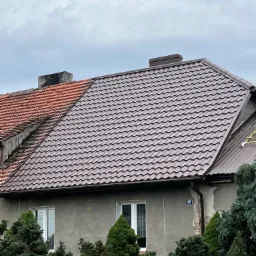 Dom z częściowo wymienionym dachem z brązowej blachodachówki, kontrastującym z fragmentem starej, czerwonej dachówki; widoczne okna i zieleń przed budynkiem.