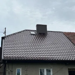Dom z nowym, brązowym dachem z dachówki ceramicznej, widoczne okno dachowe i komin, szary budynek z białymi oknami, pochmurne niebo w tle.