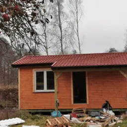 Niedokończona drewniana altana z czerwoną dachówką, widoczna osoba na drabinie i porozrzucane materiały budowlane w otoczeniu jesiennego krajobrazu.