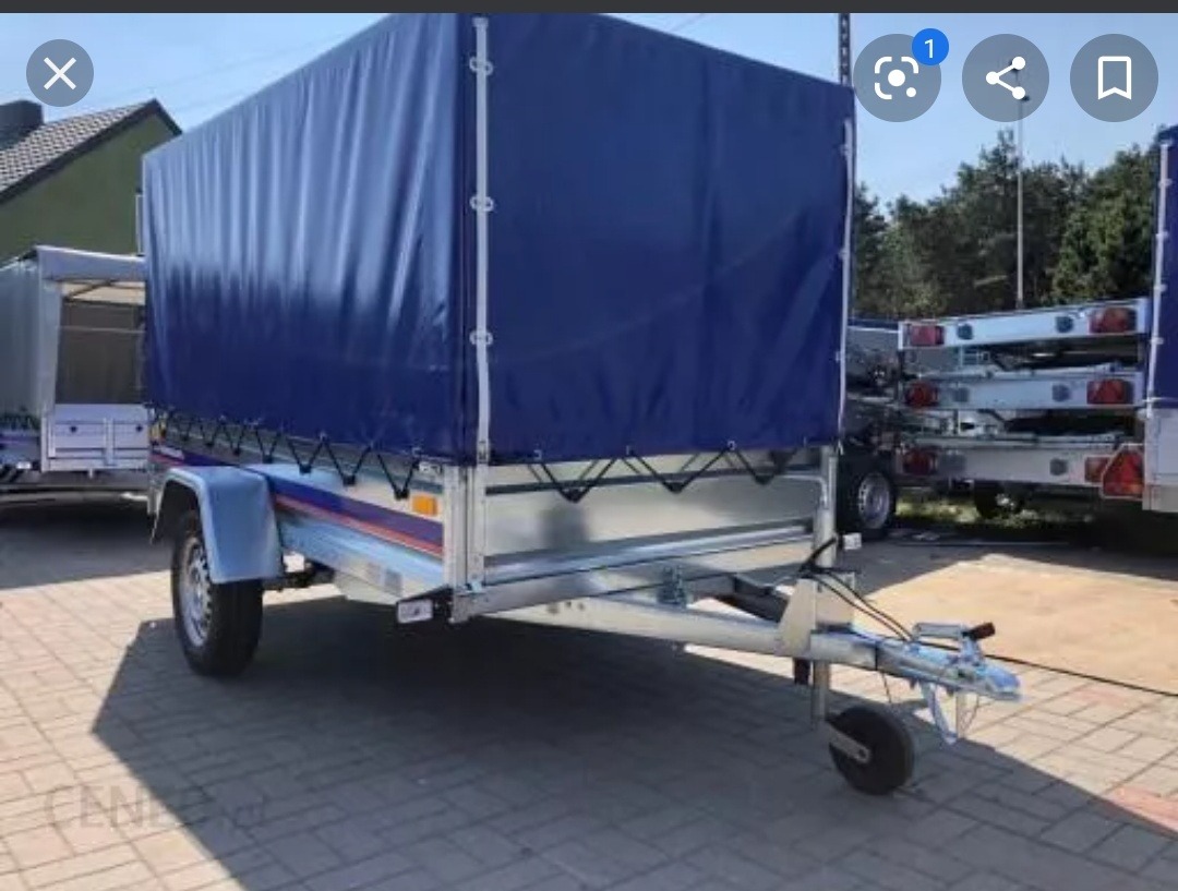 Przyczepa towarowa z plandeką w kolorze granatowym, zaparkowana na brukowanej powierzchni, z widocznymi innymi przyczepami w tle.