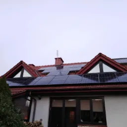 Panele słoneczne na dachu domu z czerwoną dachówką, widoczne także okna dachowe w kształcie trójkątów i komin z anteną.