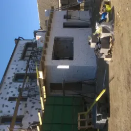 Budynek w trakcie prac elewacyjnych: ocieplanie styropianem, widoczne rusztowania, okna i otwory okienne, przygotowane bloczki styropianowe. Część budynku jest już ocieplona.