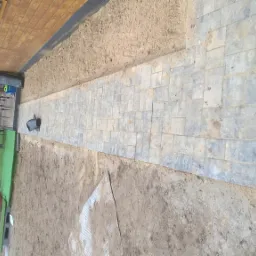 Ścieżka z szarej kostki brukowej prowadząca wzdłuż drewnianego domu, widoczna ziemia po bokach i elementy wyposażenia ogrodu w tle.