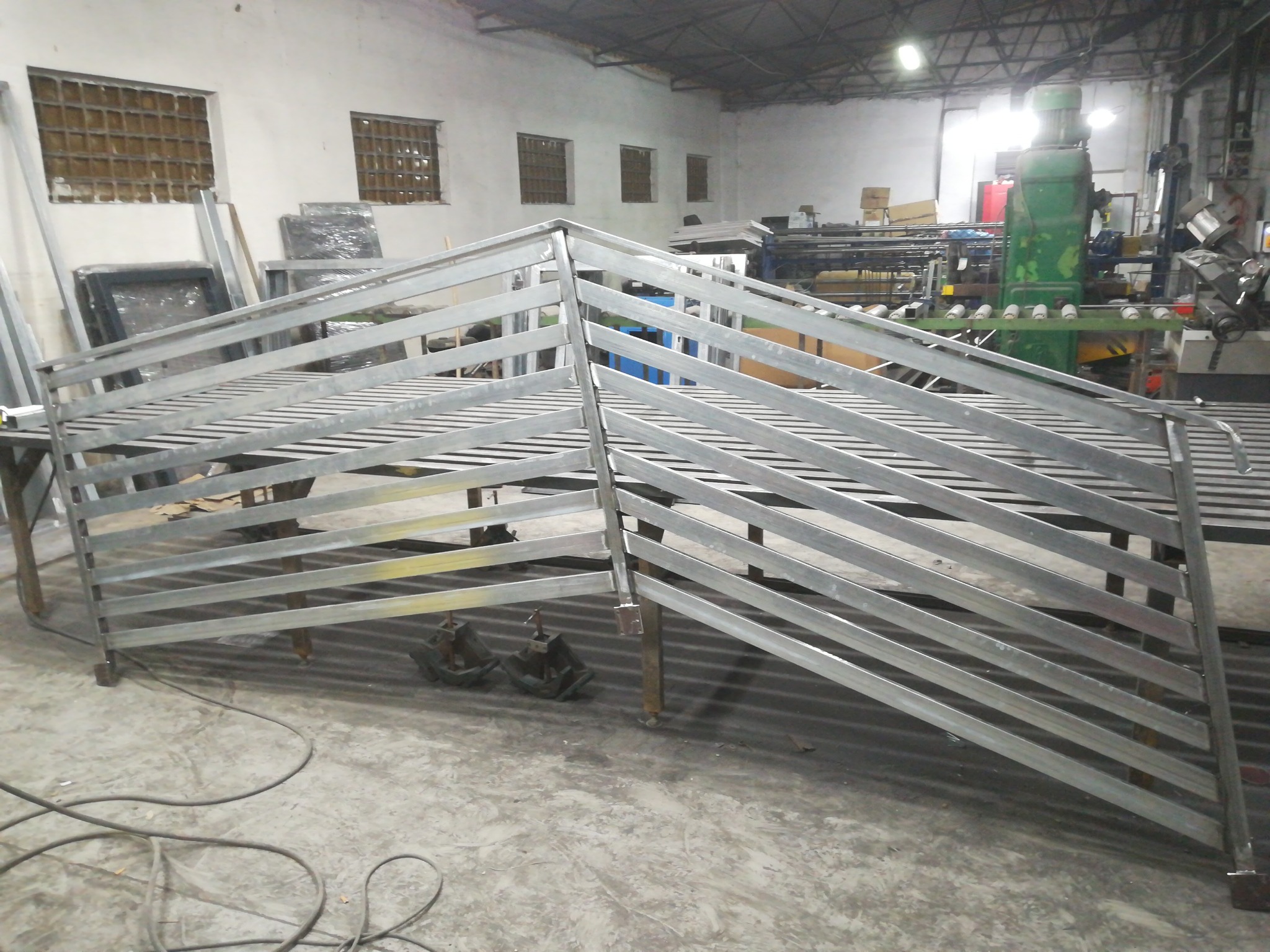 Surowa, metalowa konstrukcja balustrady lub barierki w warsztacie, widoczne elementy mocujące i spawane łączenia, w tle maszyny i narzędzia.