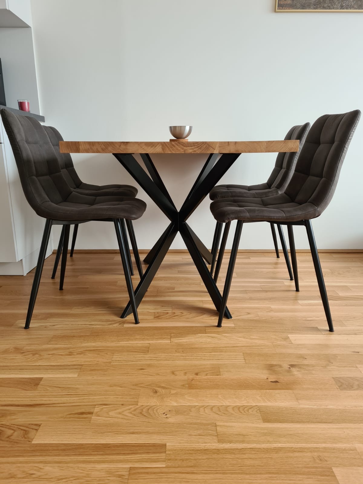 Stół z dębowym blatem i metalową podstawą w kształcie litery X, otoczony czterema tapicerowanymi krzesłami na metalowych nogach, na parkiecie.