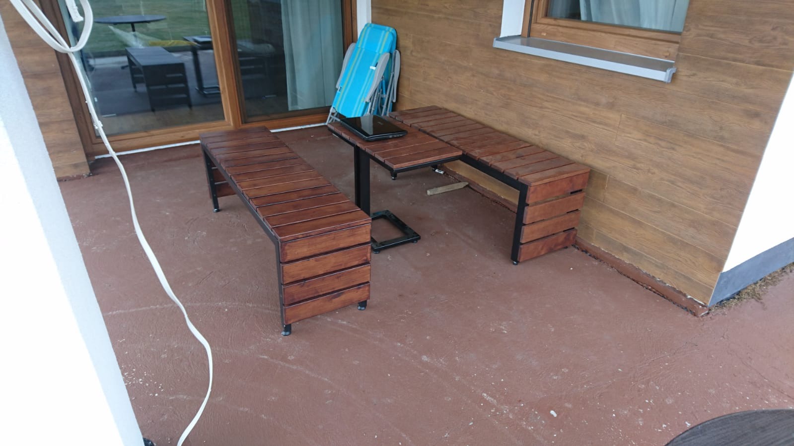 Zestaw mebli ogrodowych z drewna i metalu: dwie ławki i stół z laptopem, na tarasie z brązową podłogą i drewnianą elewacją budynku.