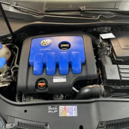 Otwarta maska samochodu, widoczny silnik TDI z niebieską pokrywą, zbiorniczki płynów i naklejki informacyjne.