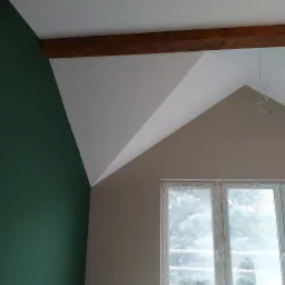 Pomieszczenie na poddaszu po świeżym malowaniu, z fragmentem zielonej ściany, białym skośnym sufitem z drewnianą belką i beżową ścianą z oknem, widoczne kable elektryczne.