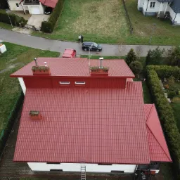 Widok z góry na dom z nową, czerwoną blachodachówką Eko-Lowicyn 3009, z widoczną drabiną opartą o ścianę i dwoma kominami, na tle zielonego ogrodu i zabudowy sąsiedzkiej.