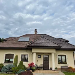 Pracownik na brązowym dachu domu jednorodzinnego z beżową elewacją, zabezpieczonym szarą plandeką, podczas mycia dachu za pomocą węża ciśnieniowego; drabina oparta o rynnę.