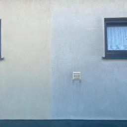 Fragment elewacji domu z widoczną różnicą w czystości powierzchni po częściowym umyciu, okno z firanką i kratka wentylacyjna.