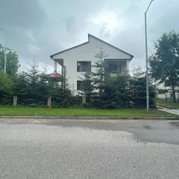 Dwupiętrowy dom z białymi ścianami i czerwonym dachem balkonu, częściowo zasłonięty rzędem gęstych, zielonych świerków.