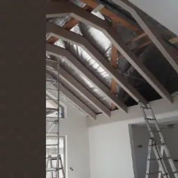 Montaż izolacji termicznej poddasza z widocznymi elementami konstrukcji drewnianej, szpachlowanymi płytami karton-gips i dwoma rozstawionymi drabinami aluminiowymi.