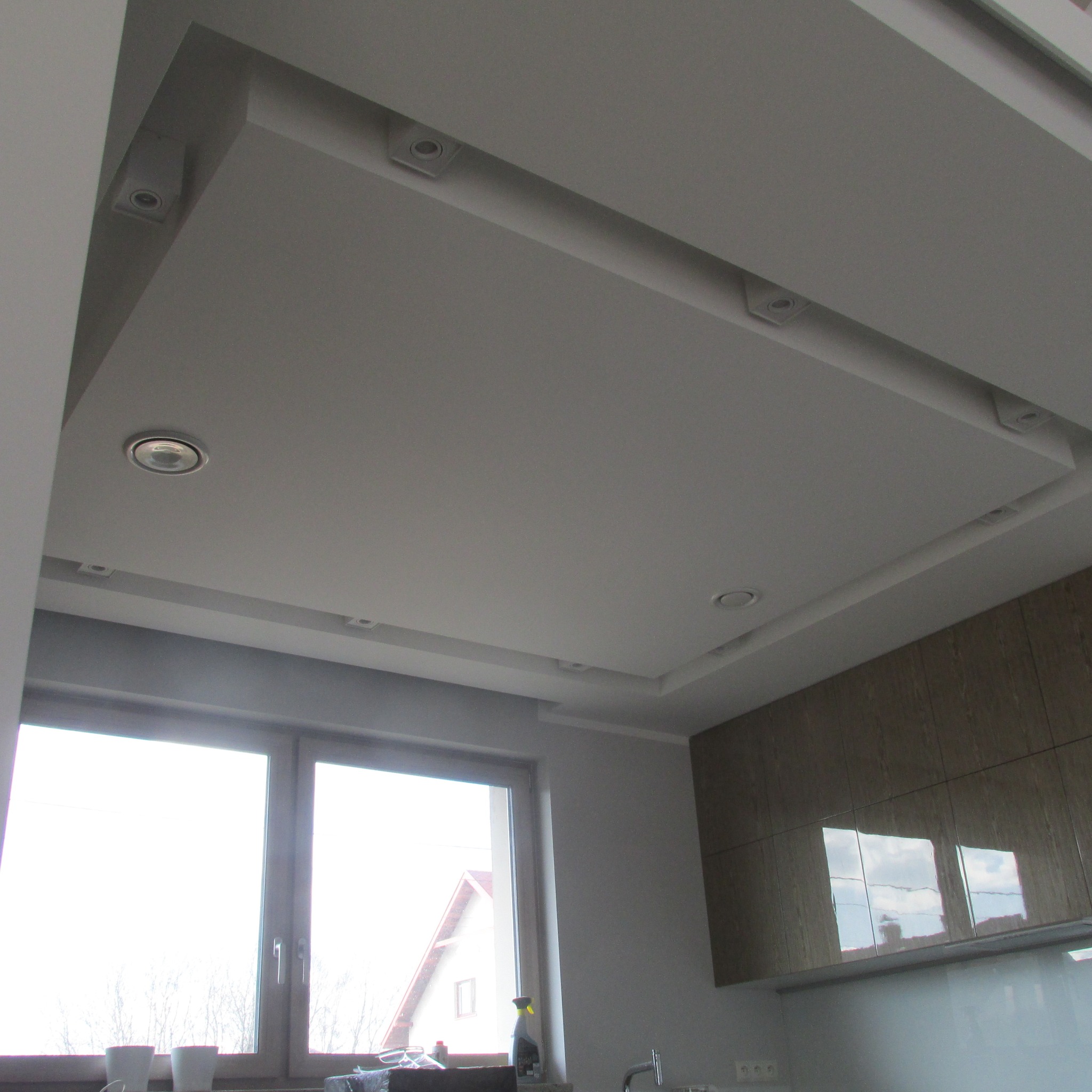 Nowoczesna kuchnia z podwieszanym sufitem z oświetleniem punktowym, jasnymi szafkami i dużym oknem.