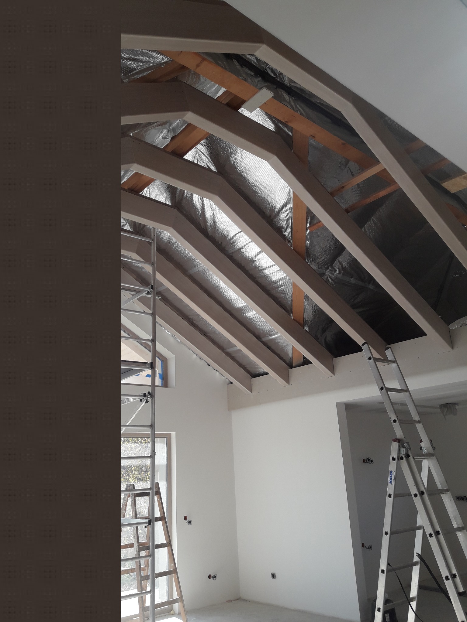 Montaż izolacji termicznej poddasza z widocznymi elementami konstrukcji drewnianej, szpachlowanymi płytami karton-gips i dwoma rozstawionymi drabinami aluminiowymi.