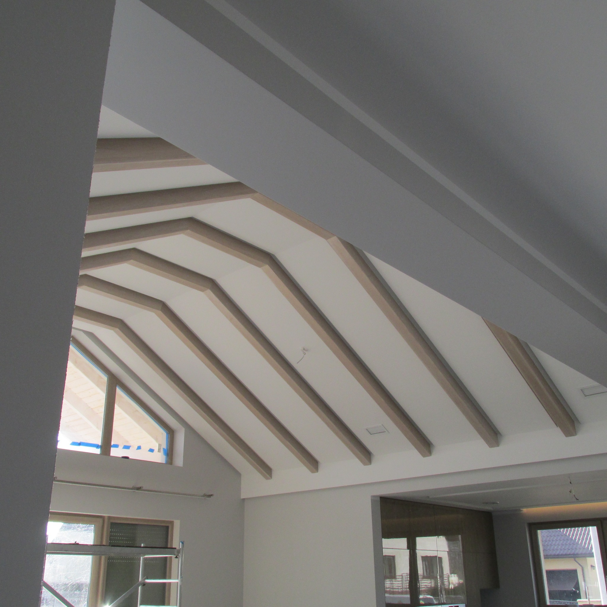 Skośny sufit z drewnianymi belkami i płytami gipsowymi, widoczne okna i fragment ściany pomalowanej na szaro.