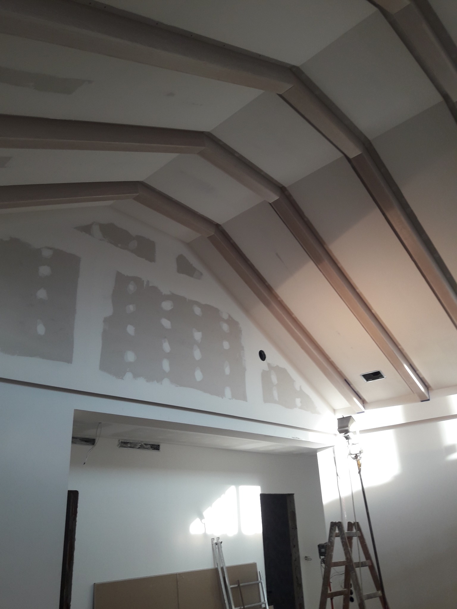 Skośny sufit z drewnianymi belkami dekoracyjnymi w trakcie montażu, widoczne ślady po szpachlowaniu na ścianie, drabina rozstawiona w pomieszczeniu.