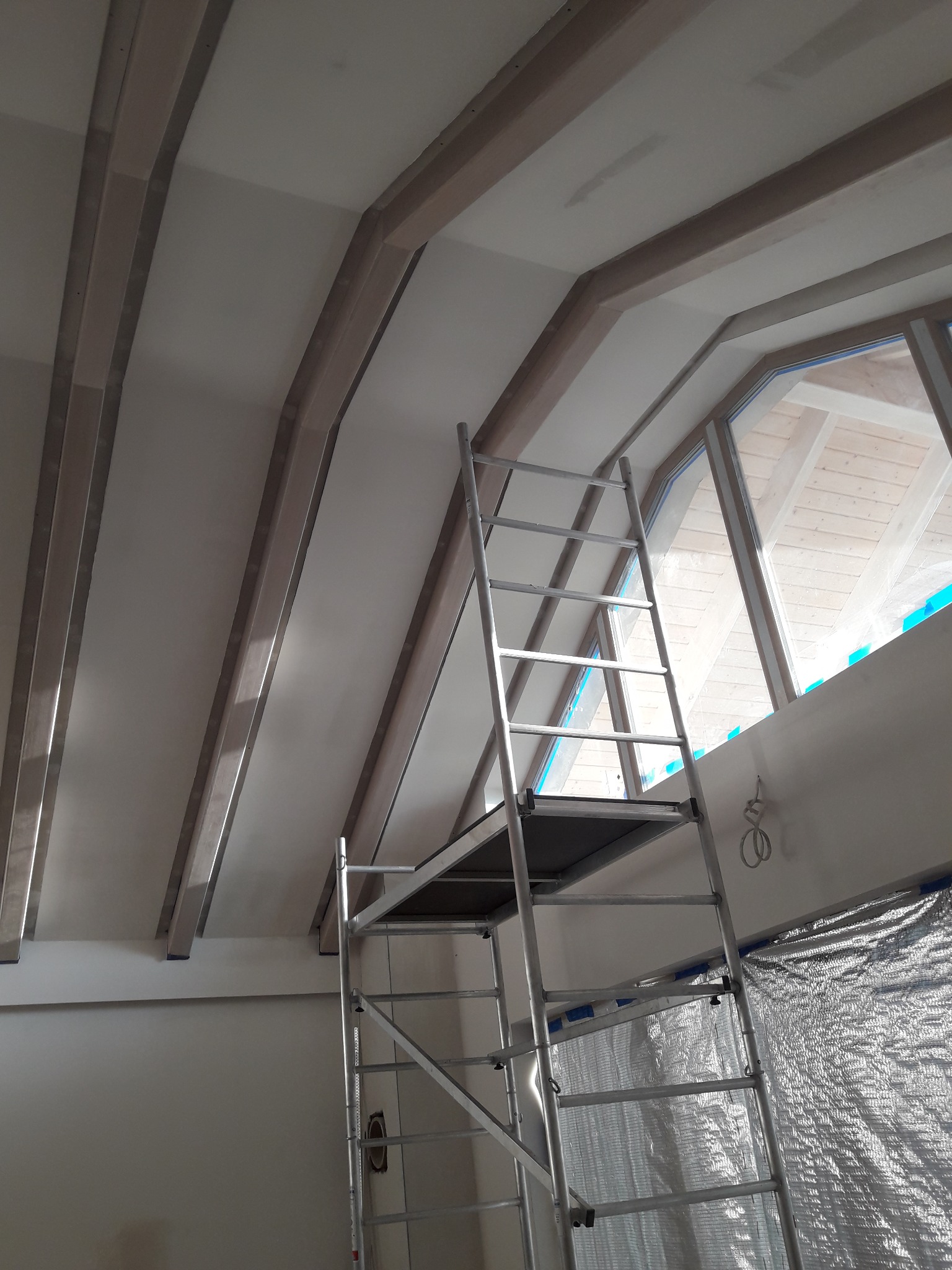 Sufit poddasza z drewnianymi belkami konstrukcyjnymi i oknem dachowym w trakcie szpachlowania, widoczny aluminiowy ruszt.