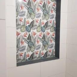 Wnęka w ścianie wyłożona płytkami z motywem tropikalnych liści i kwiatów rajskiego ptaka, otoczona ciemnoszarą ramą i jasnymi, gładkimi płytkami ściennymi.