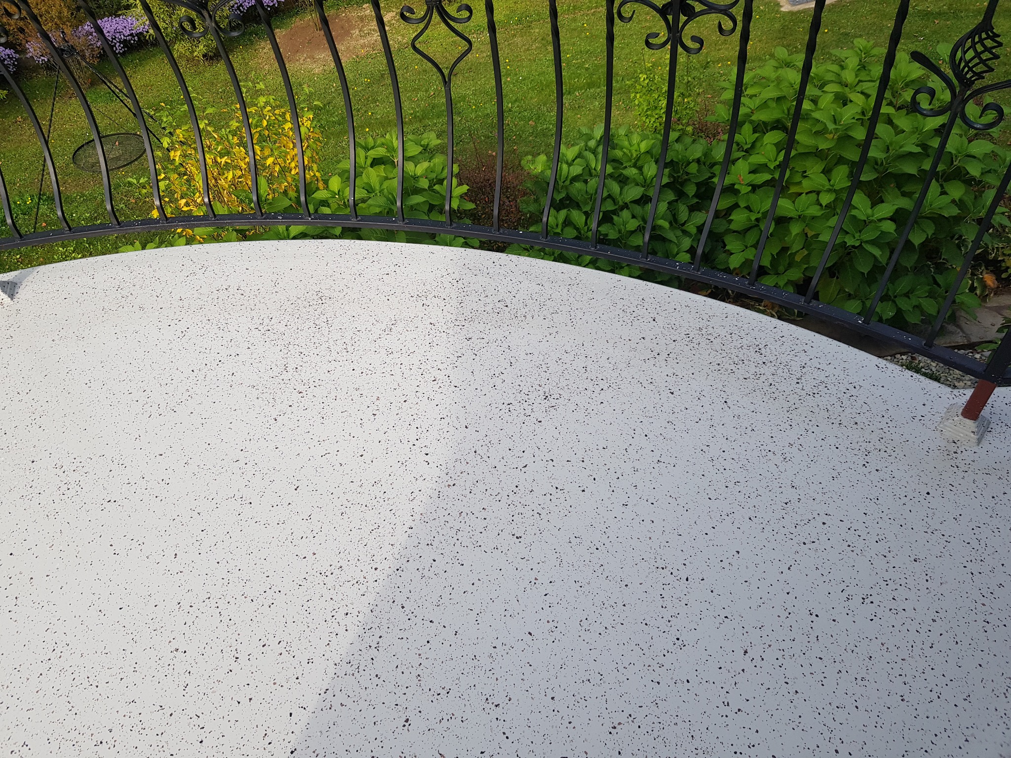 Biały, świeżo wykończony balkon z widokiem na zielony ogród przez czarną, ozdobną balustradę.