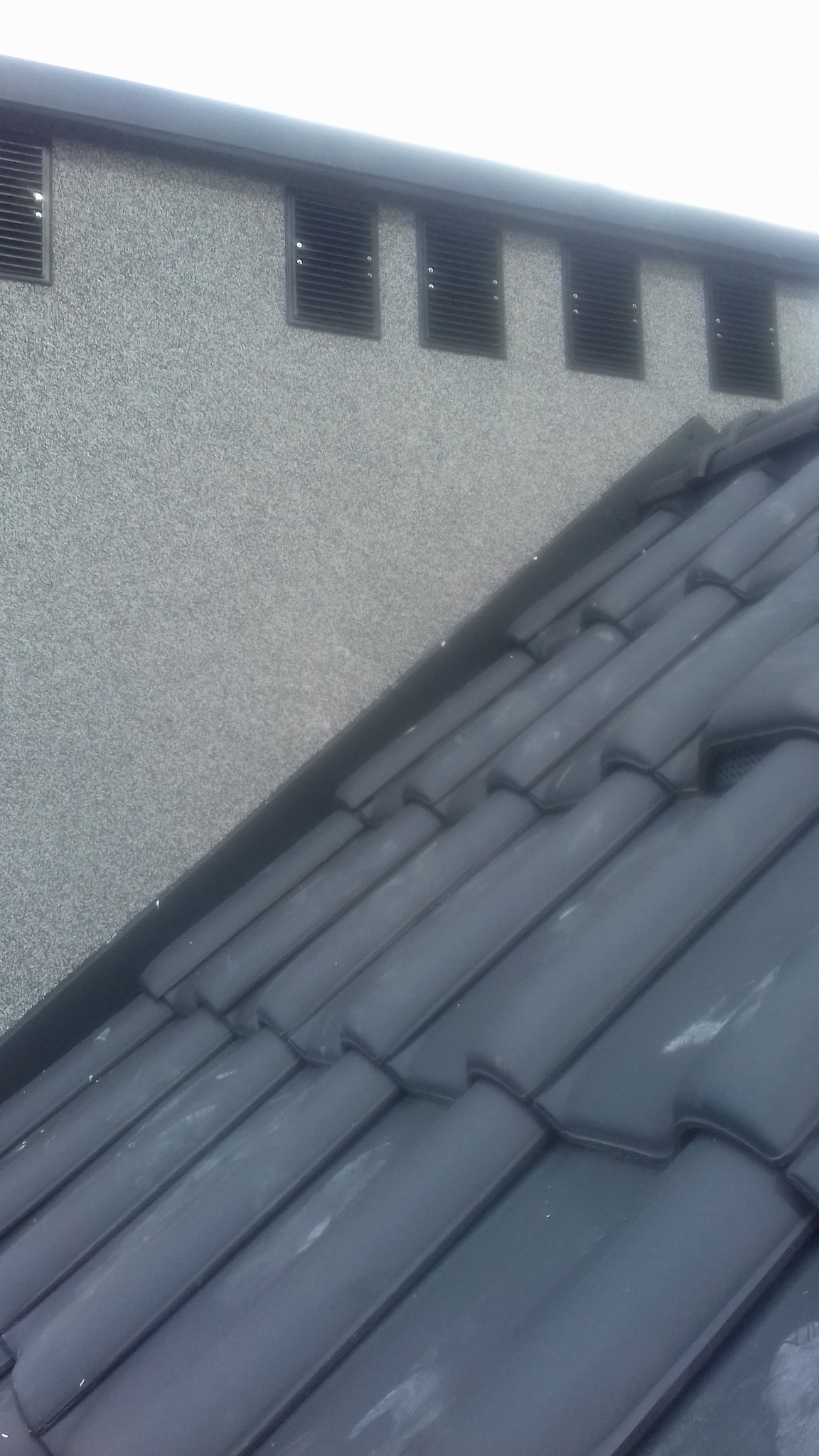 Fragment elewacji budynku z szarą, ziarnistą strukturą oraz fragment dachu pokrytego ciemnymi dachówkami ceramicznymi.