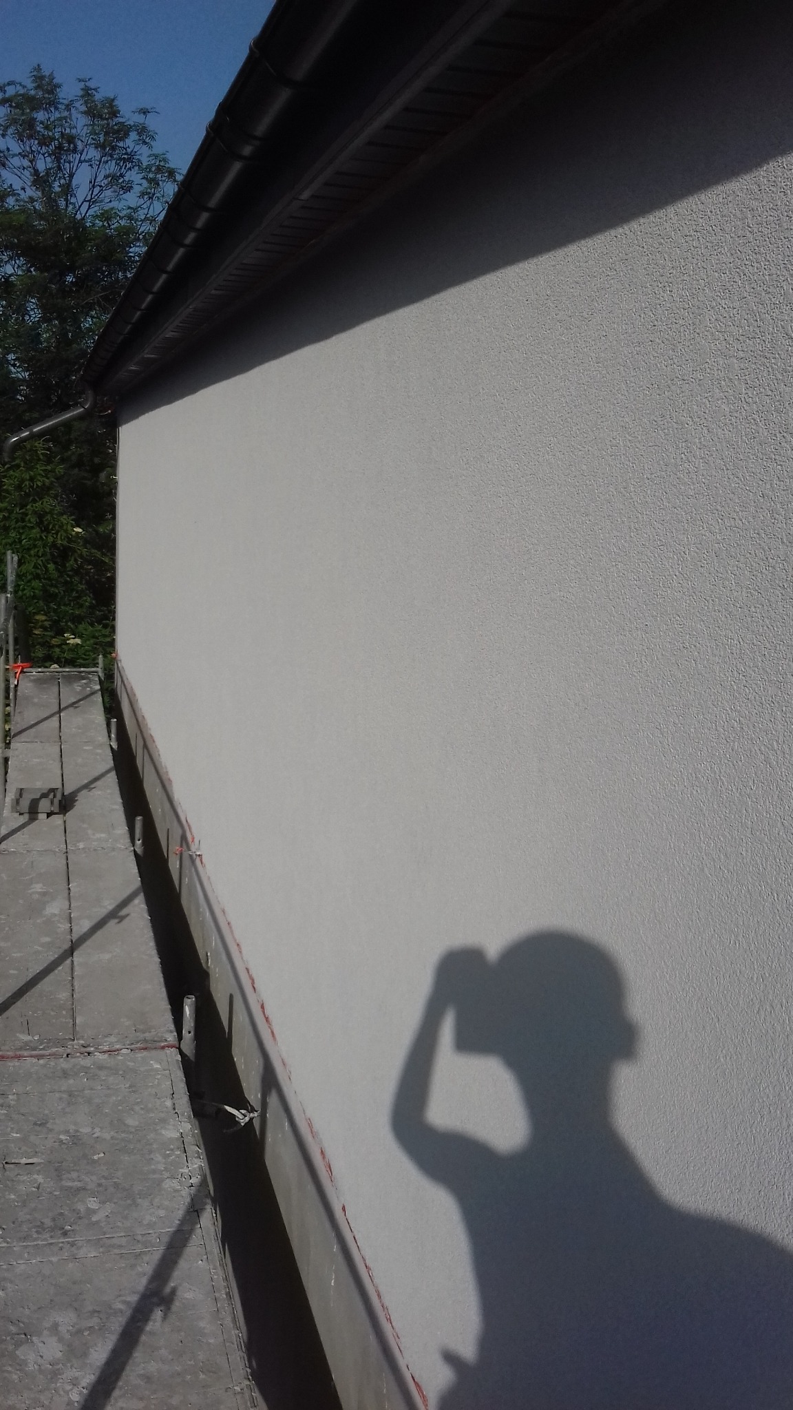 Świeżo otynkowana elewacja domu jednorodzinnego, widoczna z rusztowania, z cieniem osoby robiącej zdjęcie.