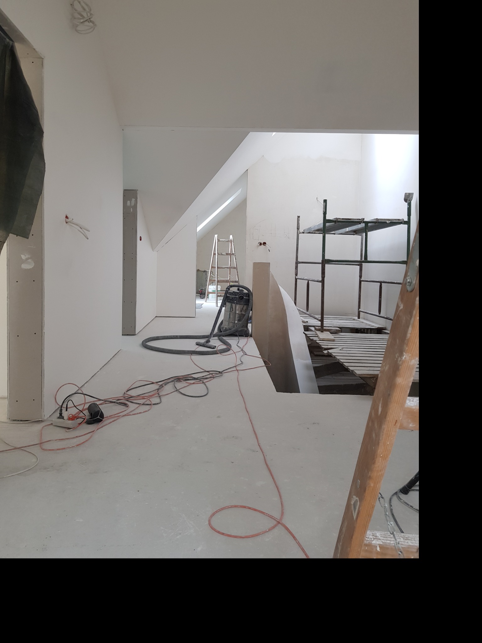 Wnętrze w trakcie wykańczania: widoczne ściany przygotowane do malowania, drabiny, rusztowanie, odkurzacz przemysłowy i rozłożone kable elektryczne.