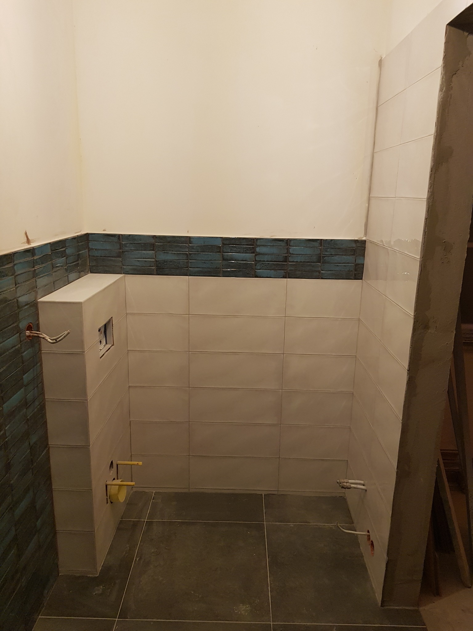 Wnętrze łazienki w trakcie remontu, widoczne ułożone płytki ceramiczne w dwóch kolorach: białe prostokątne oraz wąskie, niebiesko-szare, instalacja wodna i elektryczna wyprowadzona ze ściany...