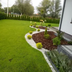 Ogród przydomowy z równo przystrzyżonym trawnikiem, ozdobnymi krzewami ułożonymi wzdłuż ścieżki z białego kamienia, oraz rabatą wysypaną korą i obsadzoną iglakami.