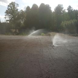 TULIPANEK Usługi Ogrodnicze - Automatyczne nawadnianie nowo przygotowanego terenu pod trawnik, z widocznymi zraszaczami i wilgotną glebą, drzewa w tle.