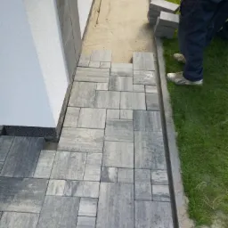 Układanie szarej kostki brukowej przy ścianie budynku z kranem, widoczne przygotowane podłoże i trawnik.