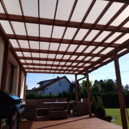 Drewniana pergola z przezroczystym zadaszeniem nad tarasem z meblami ogrodowymi, grillem i widokiem na dom z ogrodem.