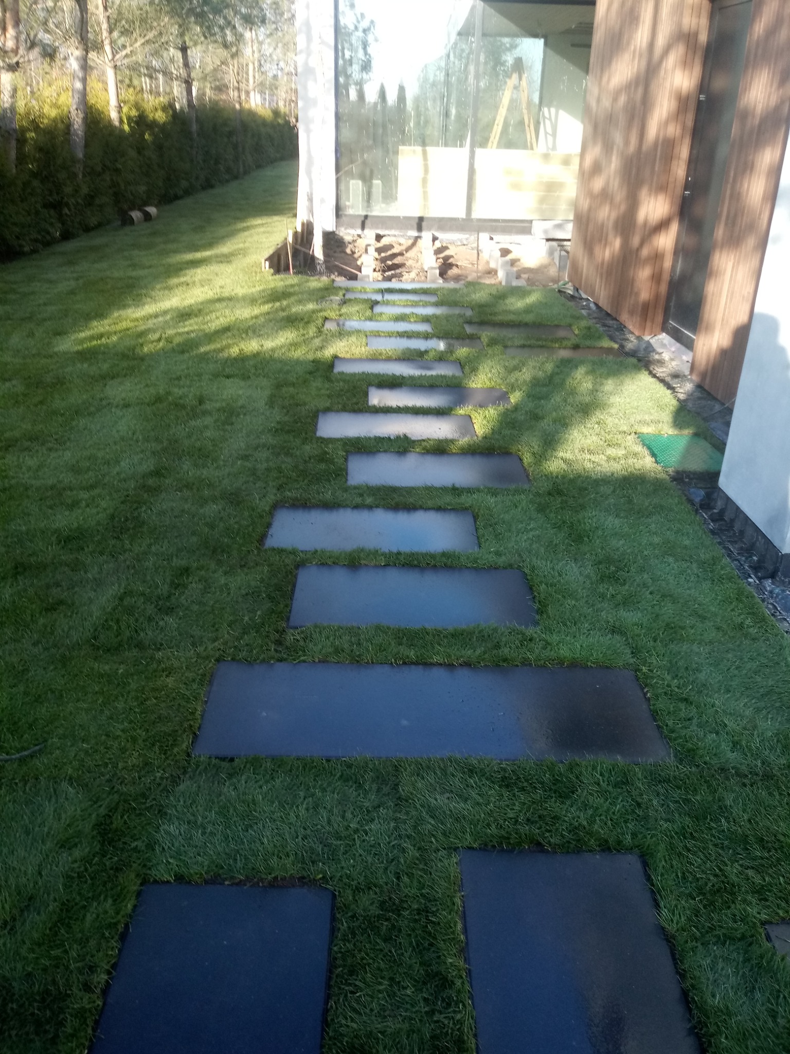 Świeżo ułożony trawnik z rolki z elegancką ścieżką z ciemnych, prostokątnych płyt prowadzącą wzdłuż nowoczesnego domu z dużymi oknami.