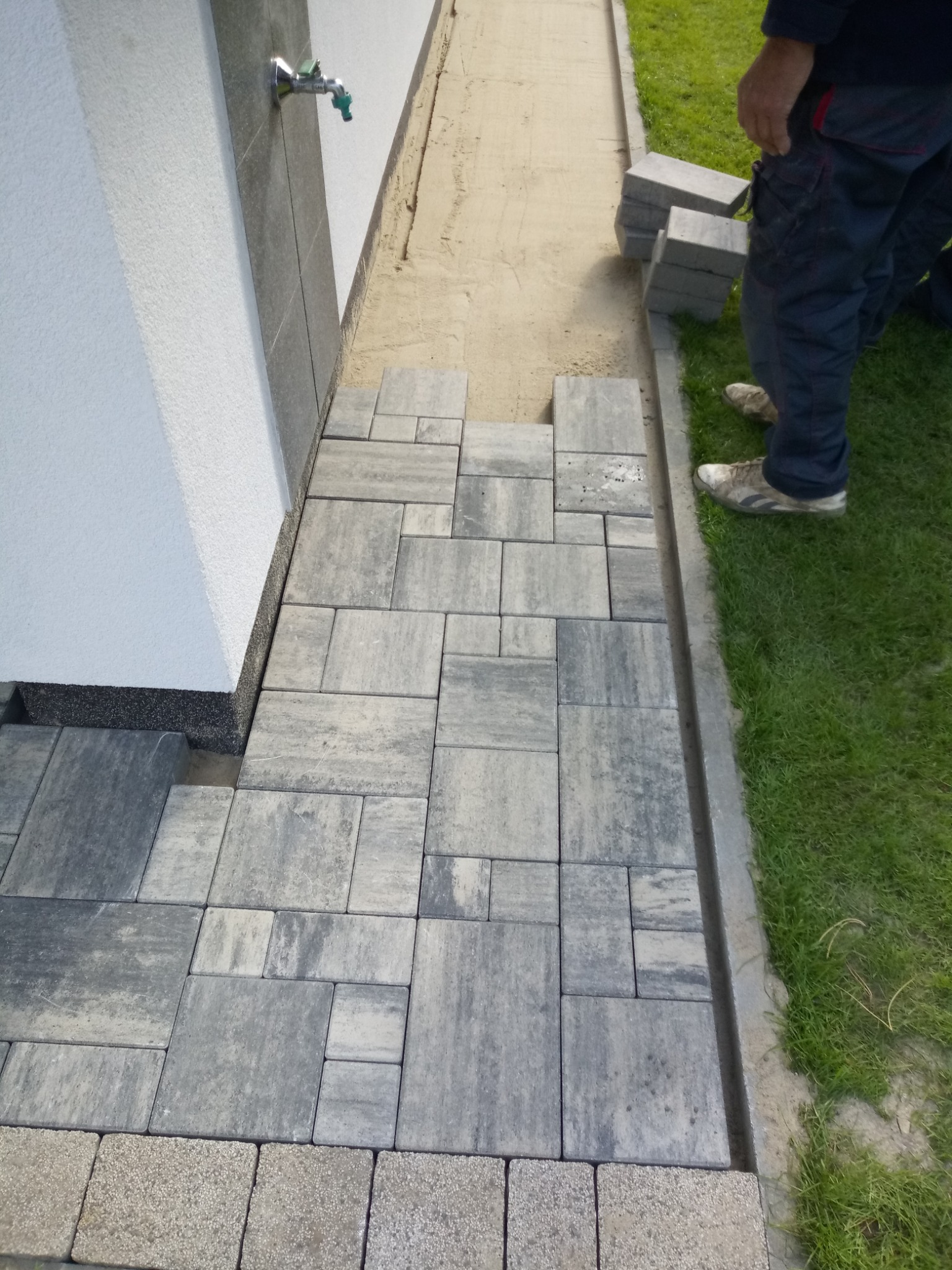 Układanie szarej kostki brukowej przy ścianie budynku z kranem, widoczne przygotowane podłoże i trawnik.