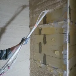 Aplikacja tynku maszynowego na ścianę z bloczków, widoczne przewody i pistolet natryskowy, fragment ściany już pokryty tynkiem, ślady spoin między bloczkami.