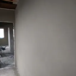 Gładka, jasnoszara ściana po szpachlowaniu, widoczna w surowym wnętrzu w trakcie remontu, z fragmentem drewnianej konstrukcji stropu i widokiem na kolejne pomieszczenie w tle.