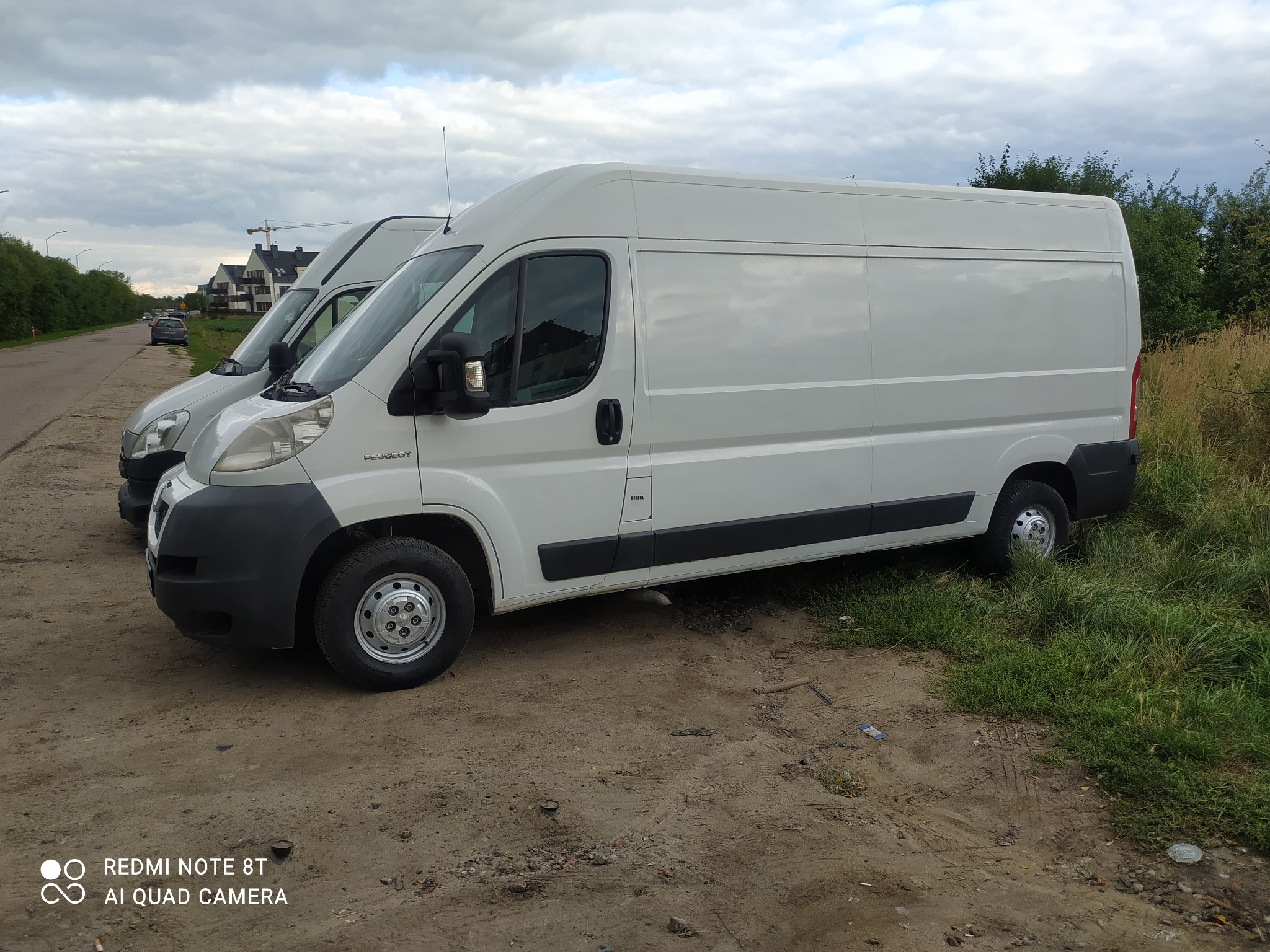 Biały samochód dostawczy Peugeot Boxer zaparkowany na szutrowym parkingu obok zielonej trawy, z drugim busem widocznym w tle i pochmurnym niebem.
