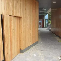 Wykończenie elewacji budynku drewnianą boazerią pionową, z widocznymi detalami łączeń i szarą podmurówką, na tle brukowanej ścieżki prowadzącej do przeszklonego wejścia.